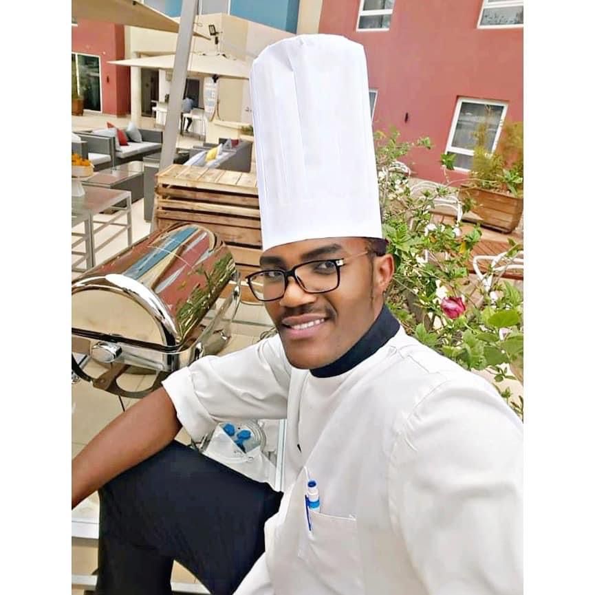 Chef Emmanuel TUYISHIME wearing a white chef hat and long cooking coat, preparing food in Lewiston Maine kitchen