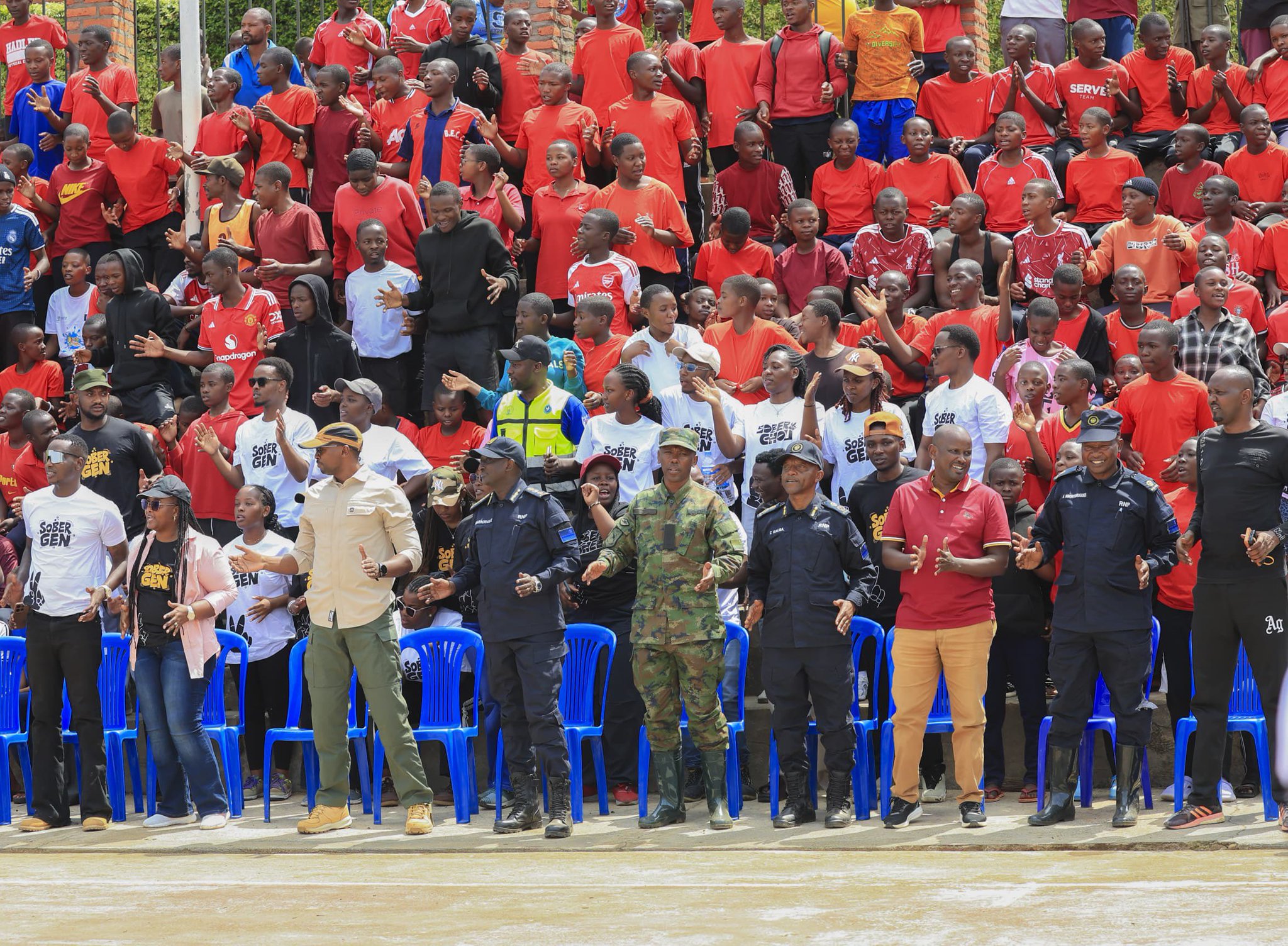Health Minister Dr. Sabin Nsanzimana and Inspector General of Police CG Felix Namuhoranye, with Rwanda Defence Force security forces and youth, standing and clapping during the SOBER GEN campaign against drug abuse and illicit alcohol in Rwanda. The crowd watches an artist performing at the event, though the artist is not visible in this photo.