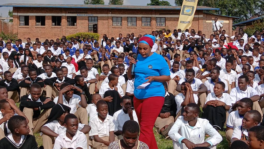 The Fight Against Drug Abuse: Marie Claire Uwineza, also known as MUTIMAWURUGO Claire, speaks to a large group of school students wearing uniforms on a sunny day in Rwanda. She holds a microphone and shares a message against drug abuse. The students listen attentively. The scene shows youth engagement, school awareness, and the fight against drug abuse in Rwanda.
